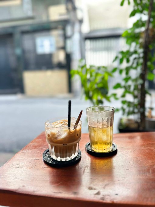 Two drinks, one iced coffee and one tea, sit on coasters on a worn, reddish table beside a plant with a blurred street view in the background.