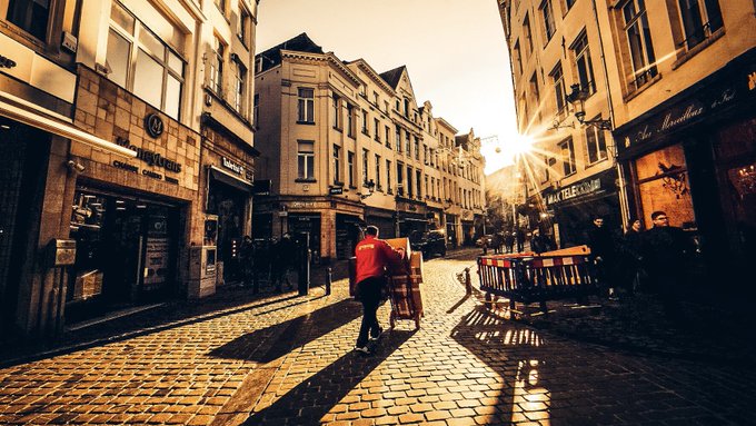 A sepia-toned city street scene at sunset with a person in a red jacket pushing a cart, sun rays peeking through buildings, and pedestrians walking on a cobblestone road.