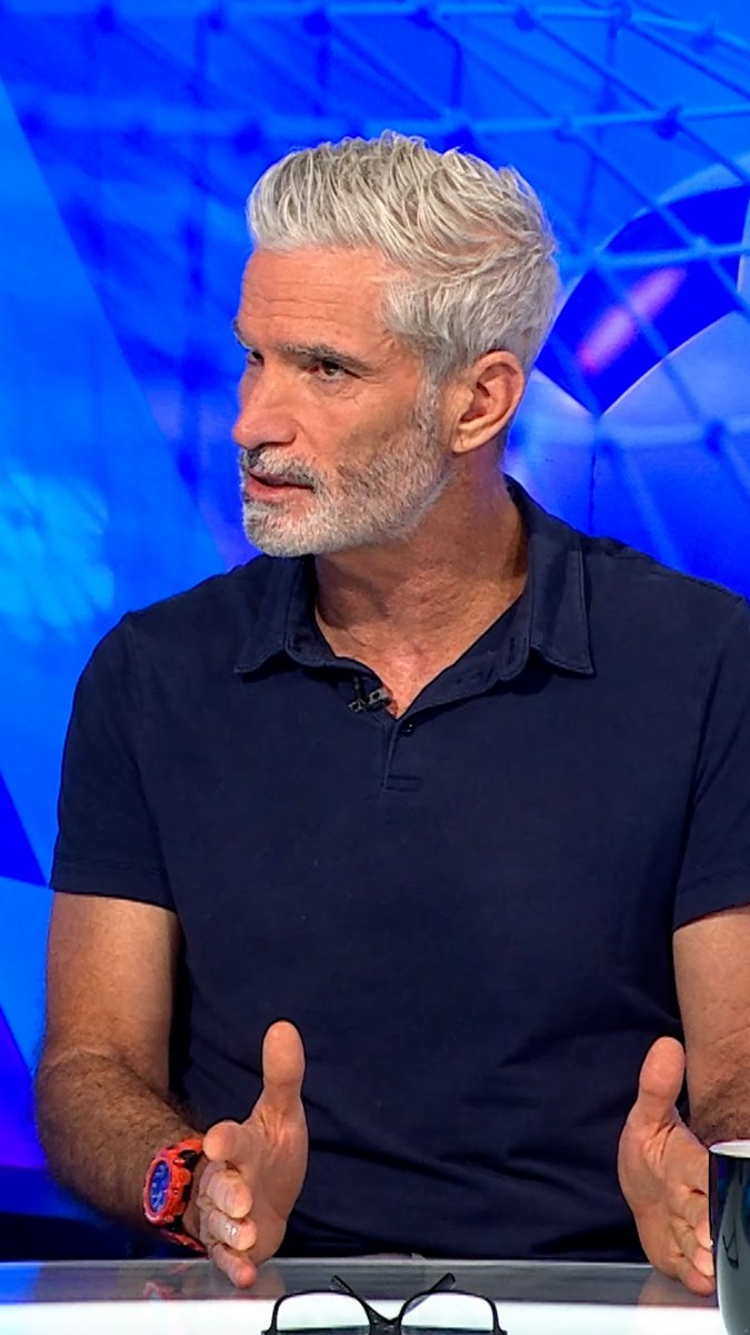 A man with styled silver-white hair and a beard, wearing a navy blue polo shirt and a red watch, is speaking while gesturing with his hands in what appears to be a television studio setting.
