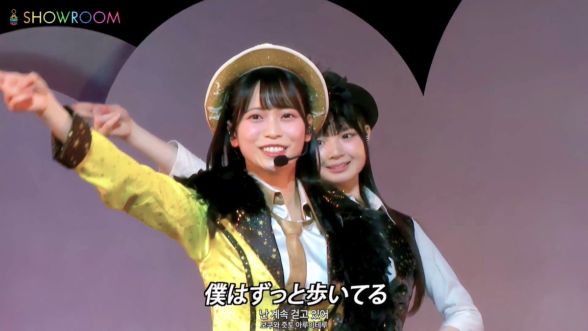 Two young female idols perform on the SHOWROOM livestream platform, with Korean subtitles reading '난 계속 걷고 있어' and Japanese text '僕はずっと歩いてる,' as they wear stage costumes with hats and star-patterned vests.