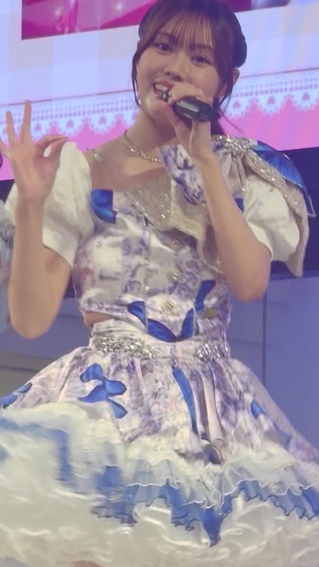 A performer in a white and blue floral patterned dress holds a microphone while smiling and making a hand gesture on stage.
