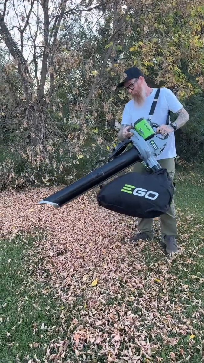 A tattooed man wearing a cap and sunglasses uses a green EGO brand backpack leaf blower, scattering a pile of autumn leaves on a lawn.