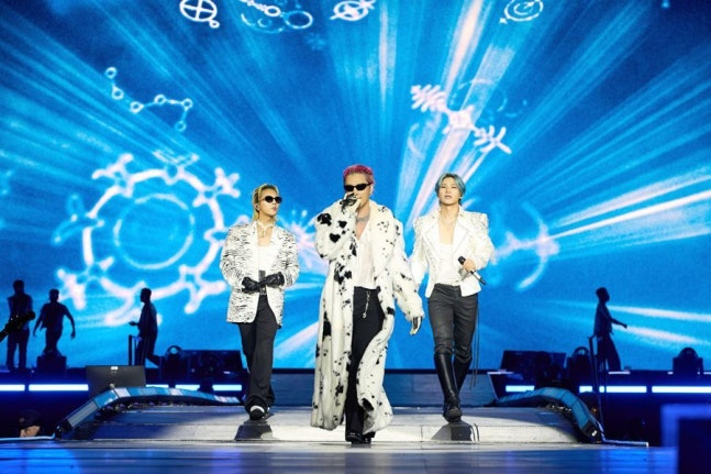 Three performers in elaborate outfits including a spotted fur coat walk forward on a stage with a vibrant blue background displaying glowing constellation and zodiac wheel graphics.