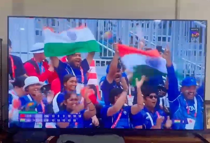 A television screen showing cricket fans in blue jerseys celebrating enthusiastically while waving Indian flags, with score graphics visible at the bottom showing a tied match.