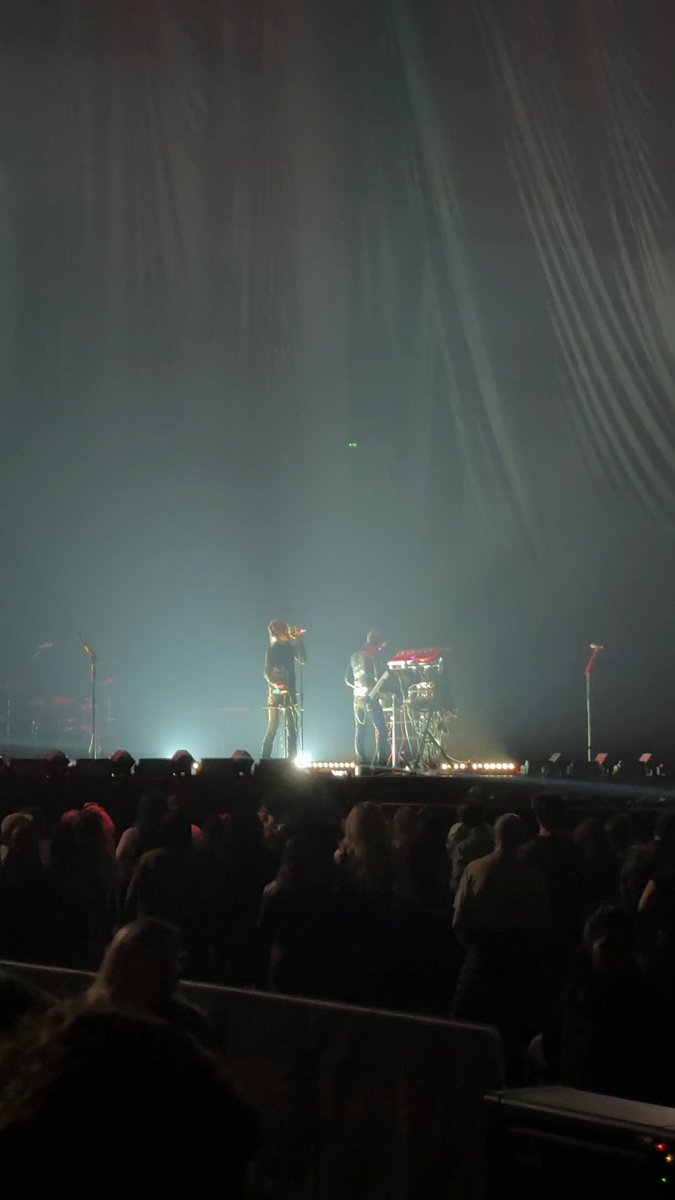 A dimly lit concert scene with two performers on stage under dramatic white spotlights, viewed from the audience.