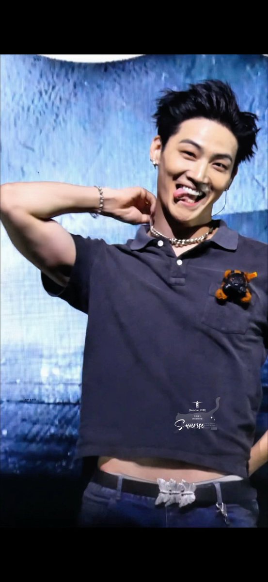 A young man with dark hair laughs joyfully while wearing a navy polo shirt with a small dog in the chest pocket, a necklace, and a belt with a flower-shaped buckle, with a 'Sunrise' watermark.