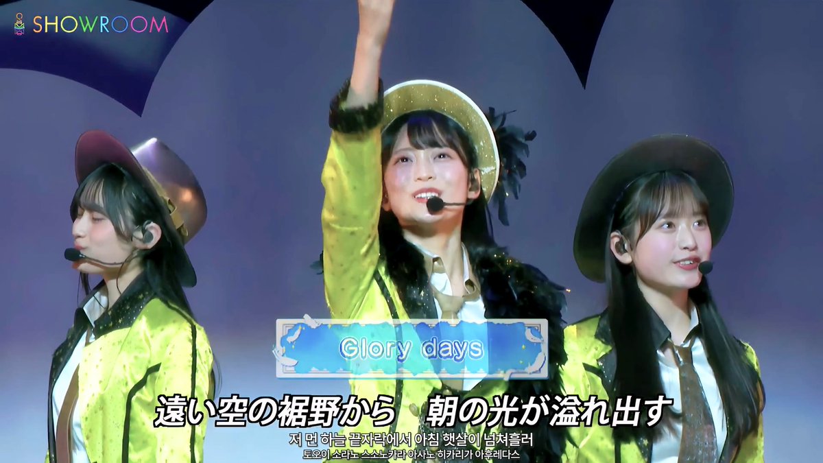 A stage performance featuring three performers in matching yellow jackets and hats, with subtitles displaying 'Glory days' in Japanese and Korean, and a 'SHOWROOM' logo.