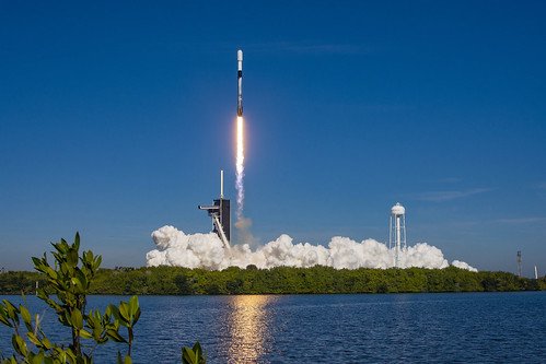 Stellarixorine's tweet photo. Soaring into a clear blue sky, a SpaceX Falcon 9 rocket thunders off the launch pad, reflected in the calm coastal waters below 🚀🌊. Massive clouds of white exhaust billow across the horizon, capturing a picture-perfect daytime liftoff set against lush Florida greenery ✨. https://t.co/KiAdjhQ0P1