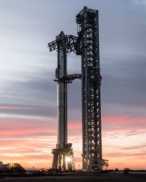 Stellarixorine's tweet photo. Fully stacked against a vibrant, soft-hued sunset 🌇, SpaceX's Starship and Super Heavy booster stand alongside their massive steel integration tower at Starbase. Held by the tower's mechanical arms 🦾, the gleaming, two-stage vehicle presents a dramatic silhouette, showcasing the immense scale and architectural ambition of next-generation space exploration.💕