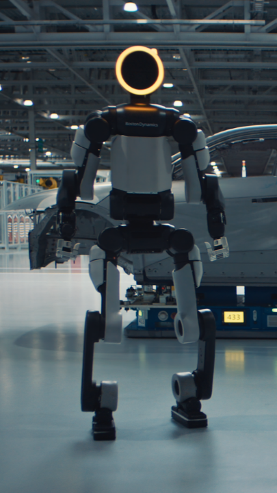 A humanoid robot stands in what appears to be a factory or workshop setting, featuring a glowing yellow ring on its head.