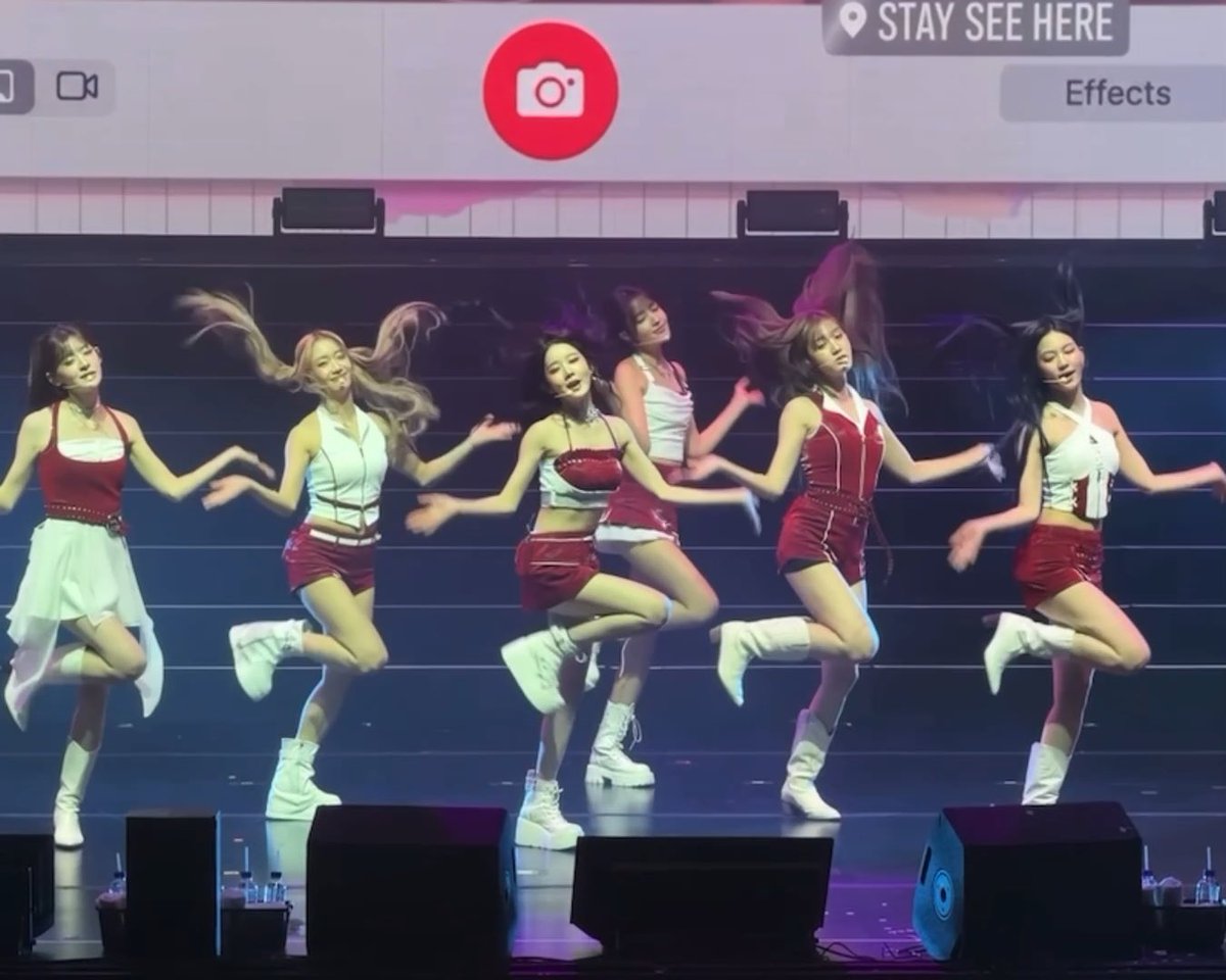 A K-pop girl group performs a synchronized dance routine on stage in matching red and white outfits with platform boots, with a camera interface visible at the top of the screen.