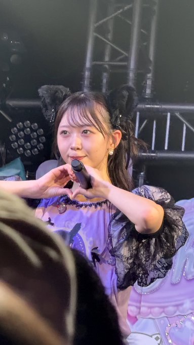 A young woman performing on stage wearing black cat ears, a purple outfit with black lace sleeves, holding a microphone and making a heart shape with her hands.