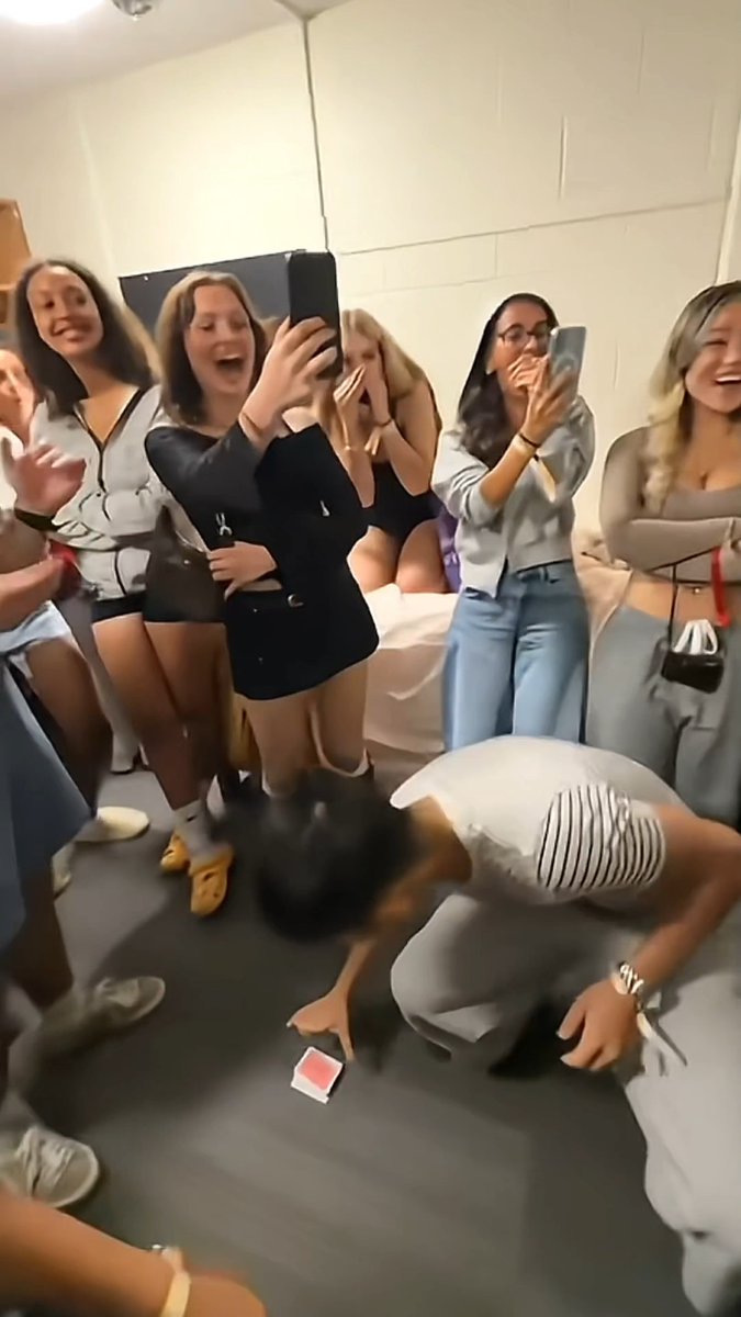 A young man performs a card trick on the floor while a group of excited young women gather around him, recording the moment on their phones in what appears to be a dorm room. The scene matches the post's humorous caption about not caring about the boy's magic ability, but rather wondering why his room is filled with so many pretty girls.