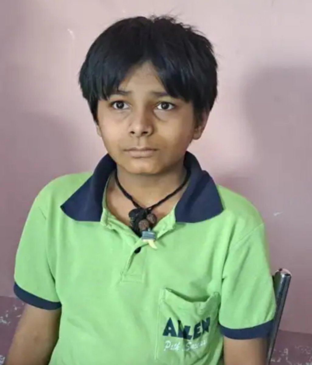 A photograph of a young boy with dark hair wearing a green polo shirt with dark blue collar and a black pendant necklace, standing against a pink wall.