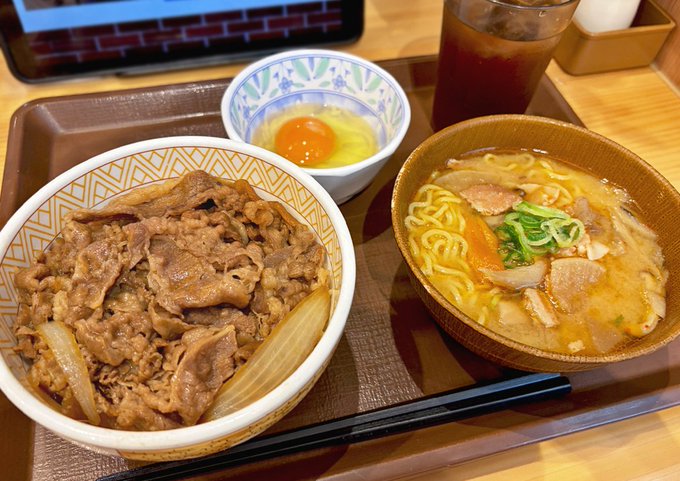 A Suka (すき家) gyudon set meal featuring a regular-sized beef bowl with onions, accompanied by tonjiru miso ramen, a raw egg, and black chopsticks on a wooden tray.