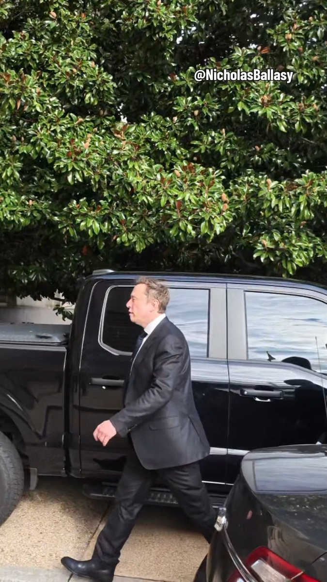 A photograph credited to @NicholasBallasy showing Elon Musk in a dark suit walking past a black pickup truck, with green trees in the background.