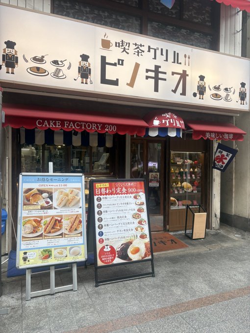 The storefront of a Japanese cafe displays signs reading 'CAKE FACTORY 200' and 'ピノキオ' (Pinocchio), with menus advertising morning and lunch options.