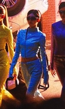 A stylized, color-filtered image of three women in form-fitting outfits (yellow, blue, and purple) and sunglasses walking confidently in what appears to be a scene from the animated series 'Arcane'.