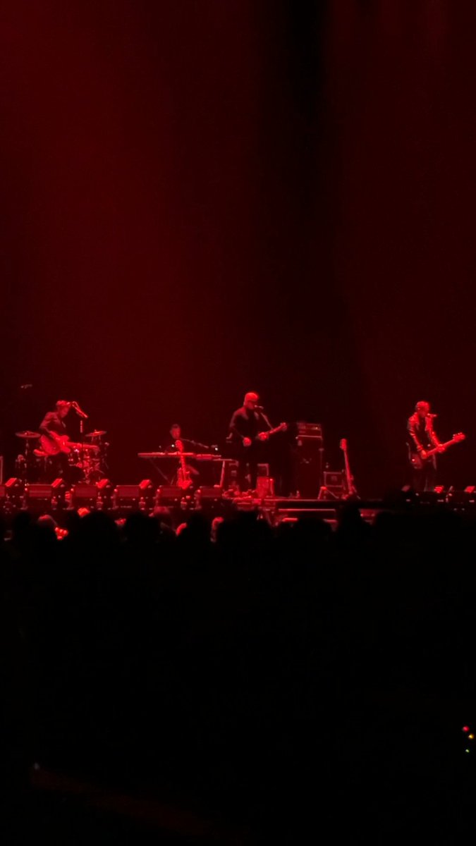 A dimly lit concert stage illuminated in red lighting, showing a band performing with guitars, drums, and keyboards in front of a silhouetted audience.