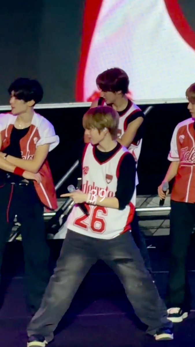 A young male performer with blonde hair dances on stage wearing a white and red "Cardinals 26" jersey, flanked by other members in matching attire.