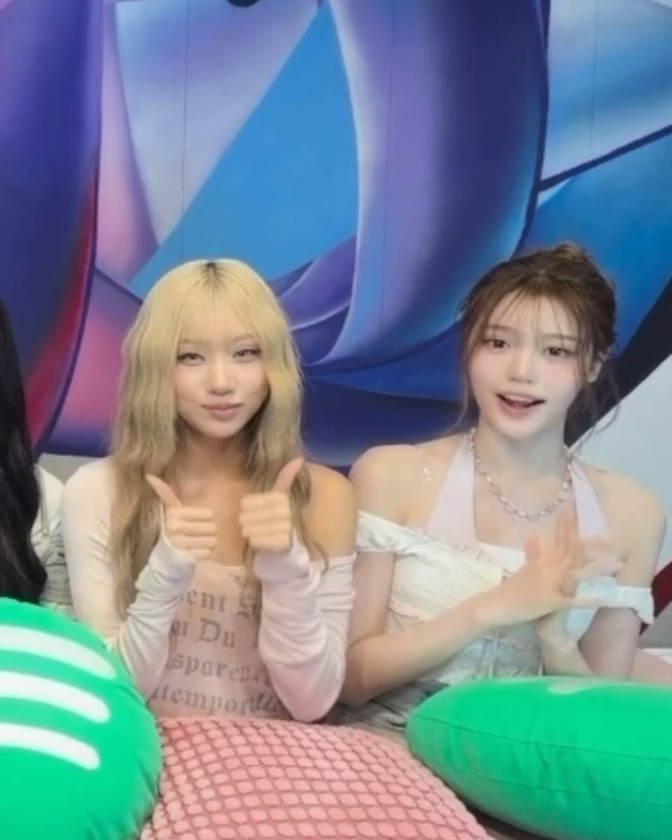 Two young women with light-colored hair pose together on a colorful set with green heart-shaped cushions, giving thumbs up and peace signs while wearing casual pink and white outfits.