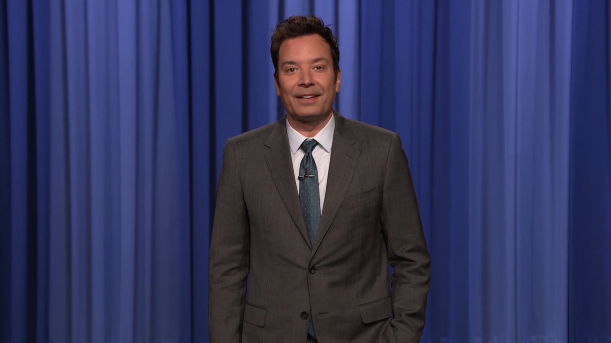 Jimmy Fallon stands on stage in a gray suit with a teal tie, smiling in front of the classic blue curtain backdrop of The Tonight Show, introducing a story about Karoline Leavitt's upcoming memoir.
