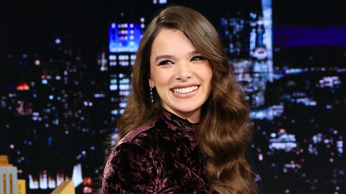 Hailee Steinfeld beams in a plum crushed velvet top on The Tonight Show, her wide smile and glamorous makeup shining against the backdrop of illuminated city skyline set pieces.

The caption aligns with the post's reference to her FallonTonight appearance, where Steinfeld is likely being welcomed back to discuss her recent marriage, new baby, and major film role.