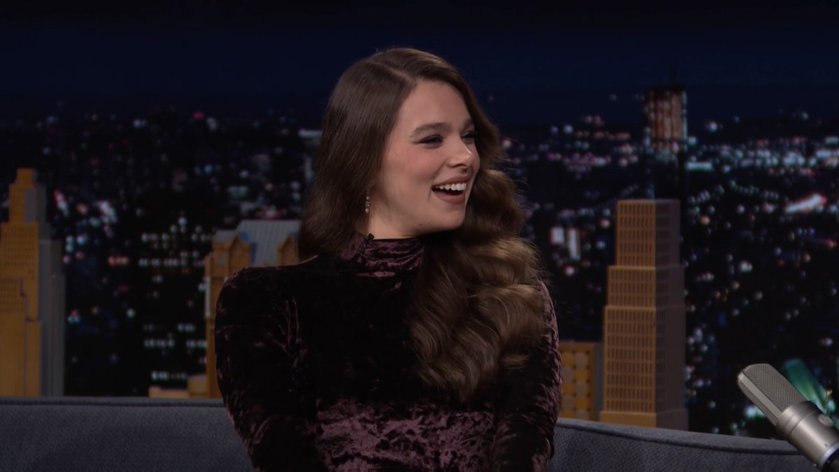 Hailee Steinfeld, wearing a dark velvet high-neck top, laughs heartily while seated on a couch during a late-night interview, with a nighttime cityscape backdrop and a silver microphone visible. The image captures her lively reaction as she addresses rumors about appearing in "Avengers: Doomsday" on "The Tonight Show."