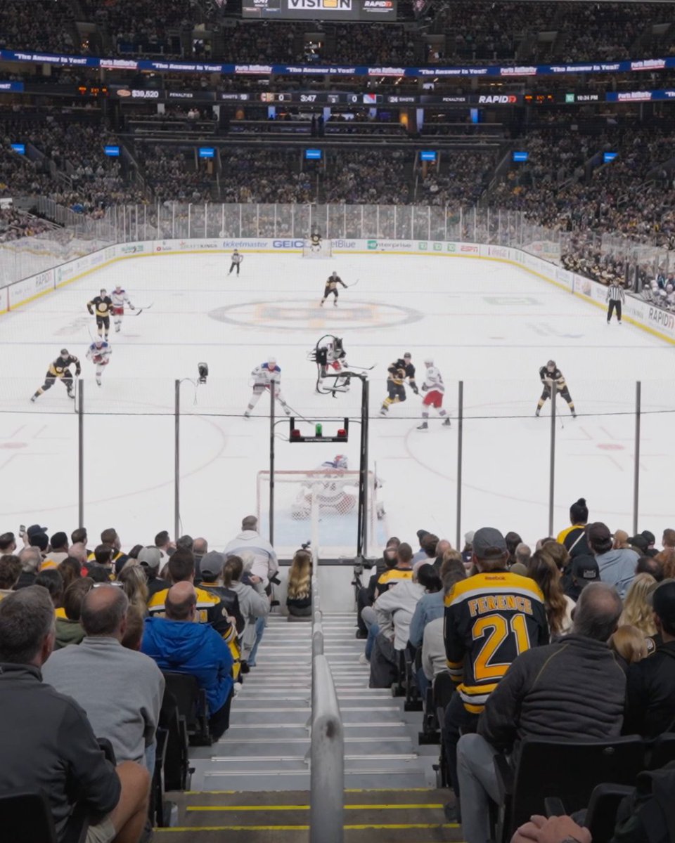 The image shows a game in progress at an NHL arena, with the scoreboard reading "Bruins 0 - Rangers 0" at 3:47 in the 1st period, while fans in the foreground wear Bruins jerseys, including one with "FRANK 21" on the back. The caption notes this is the Bruins' first goal of the season, suggesting the scoreboard will soon change from 0-0 after this matchup against the Rangers at what looks like Madison Square Garden.