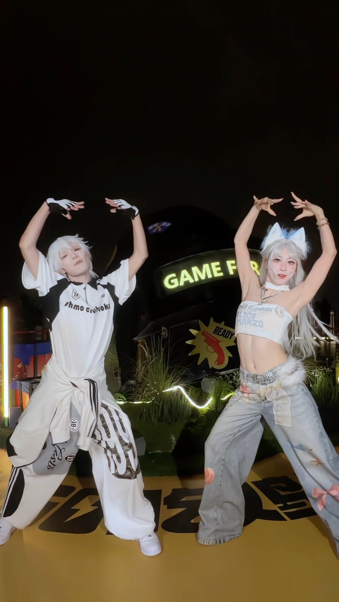 Two cosplayers pose with arms raised in a dance-like stance in front of a 'GAME FOR...' sign at night, with the person on the right wearing a white top with text including 'AMZO' and fox ears.