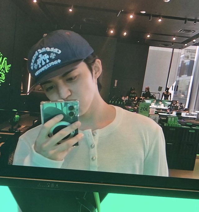 A monitor or screen displays a selfie of a young man wearing a dark baseball cap and white henley shirt, holding a smartphone while taking a mirror selfie in an electronics store.