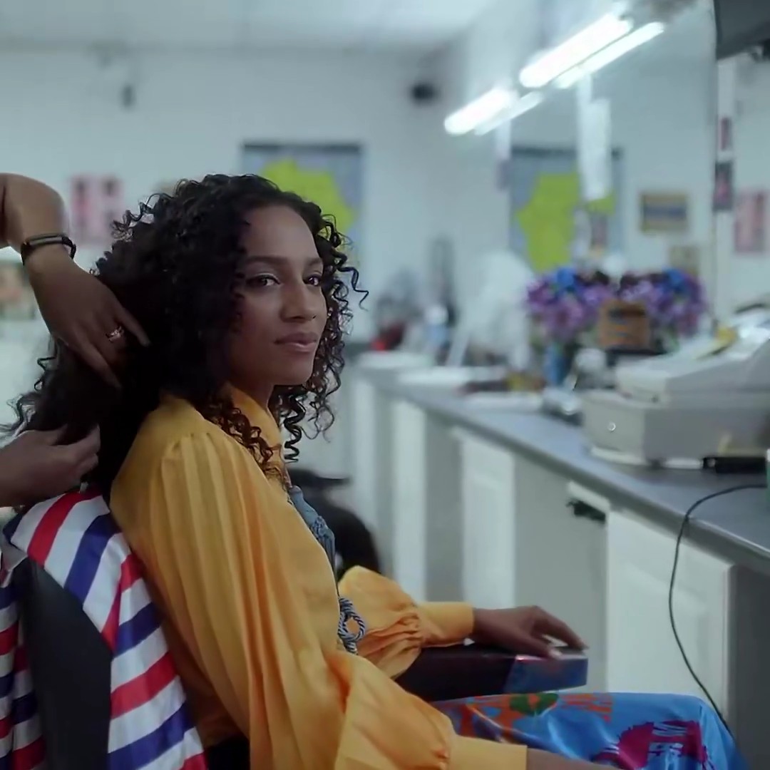 A woman with curly hair sits in a hair salon wearing a yellow blouse and colorful patterned skirt, looking over her shoulder at the camera. The image doesn't visually reference Wizkid's "Joro" anniversary but appears to be a promotional or behind-the-scenes photo shared alongside the music milestone announcement.