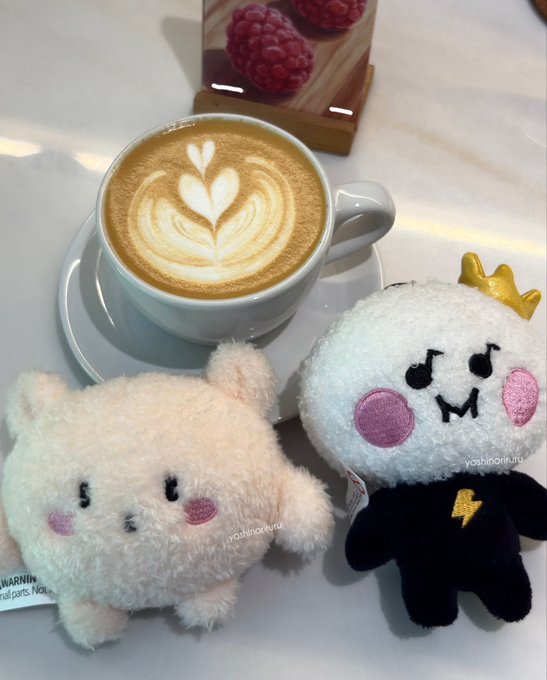 A cup of coffee with leaf-shaped latte art sits on a saucer next to two small, fluffy plush toys—one pinkish-white and one white wearing a black outfit with a gold crown.