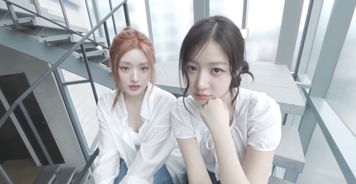 A selfie of two young women with light and dark hair, both wearing white blouses, sitting together on modern metal stairs in a bright building.