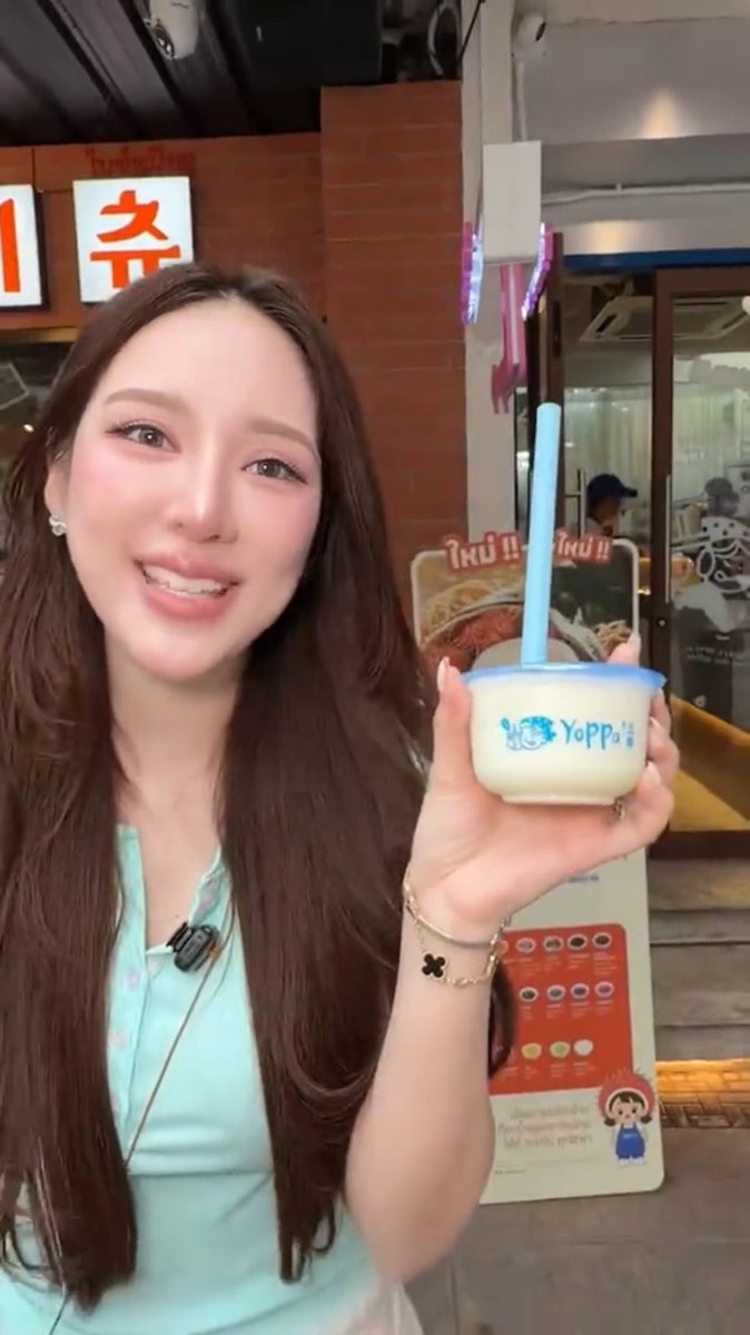 A woman with long brown hair smiles while holding a Yoppo-branded cup with a blue straw, standing in front of a shop with Korean signage.