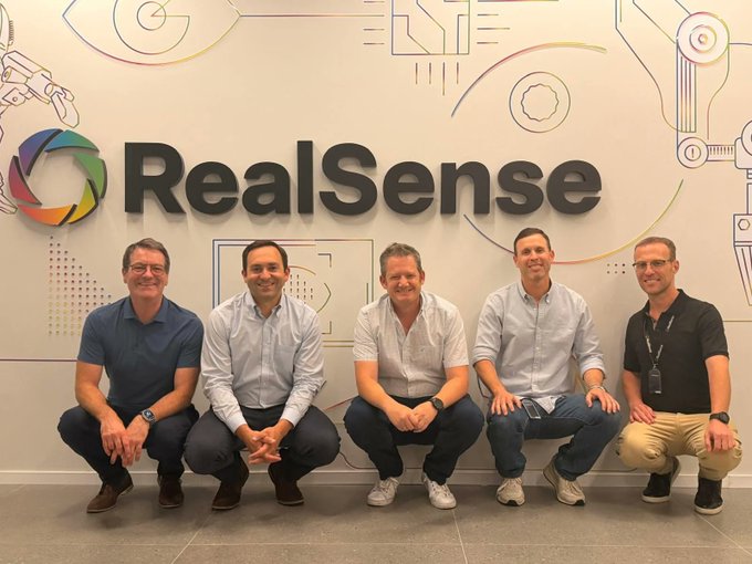 A group photo of five men in business casual attire kneeling in front of an Intel RealSense branded wall with colorful logos and technical graphics.