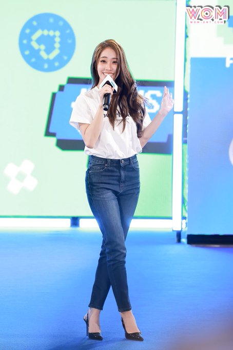 A young woman with long dark hair wearing a white shirt and blue jeans, holding a microphone and speaking at a WOM event.