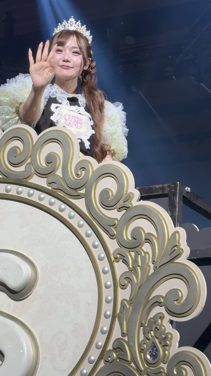 A photograph of a smiling young woman waving from an ornate golden and white carriage or float, wearing a sparkling tiara and a pastel dress with a black apron labeled 'CUE LAND'.