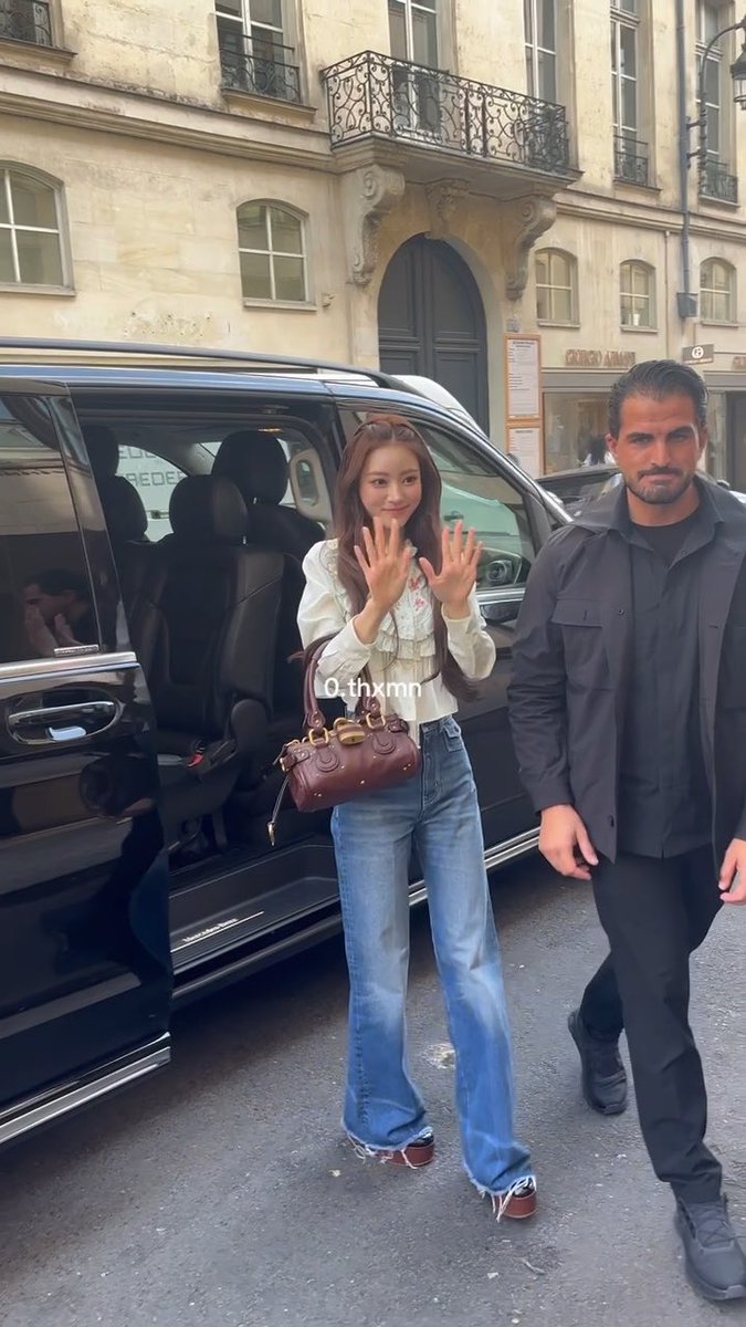 A woman in jeans and a white blouse waving next to a man in dark clothing beside a black SUV, with a 'Giorgio Armani' sign on a building in the background.