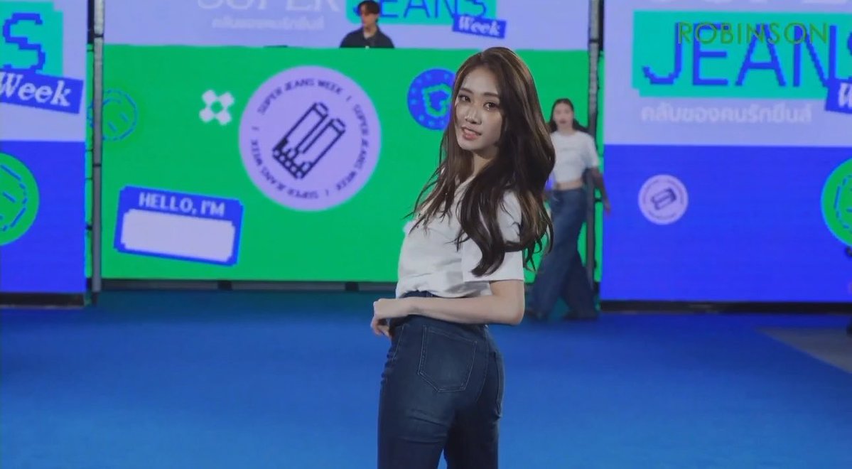 A young woman with long dark hair poses in a white t-shirt and dark jeans in front of a colorful 'ROBINSON JEANS' promotional backdrop with 'JEANS Week' branding.