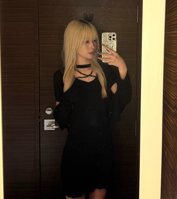A mirror selfie of a woman with long blonde hair and bangs wearing a black dress, black choker, and layered necklaces, holding a smartphone with a decorative case in front of a dark wooden door.