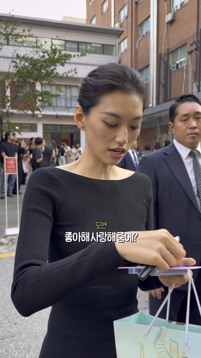 A woman in a black dress stands outdoors signing an item, with overlaid Korean text reading "도연 좋아해 사랑해좋아?" (Doyeon, do you like me? I love you, okay?), with a man in a suit standing behind her.