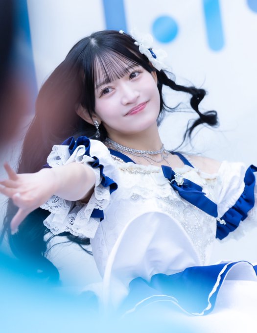 A photograph of a smiling young woman with dark hair in pigtails, wearing a frilly white and blue dress with pearl accessories and a jeweled headband, extending her hand toward the camera.