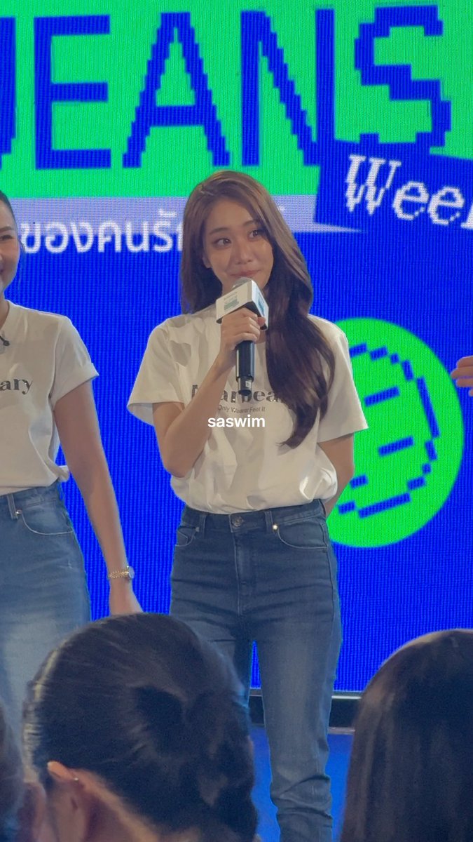A young woman with long brown hair, wearing a white t-shirt and jeans, speaks into a microphone on a stage in front of a blue and green screen displaying 'EANS Week' and 'saswim'.