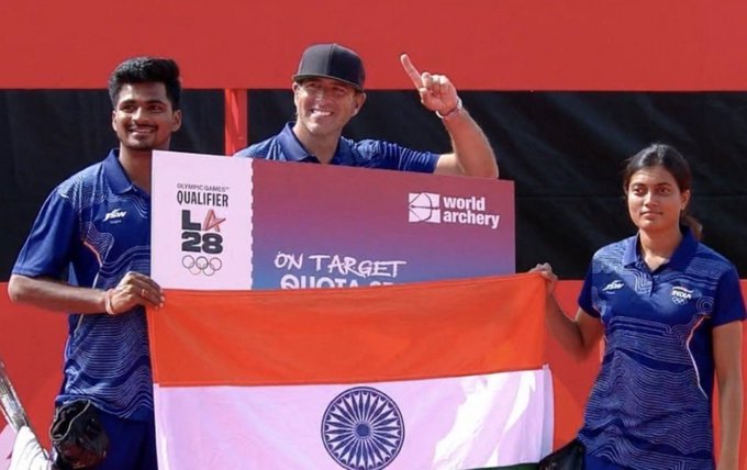 Three archers in blue uniforms posing with the Indian flag and an 'Olympic Games LA 2028 Qualifier - On Target' banner by World Archery.