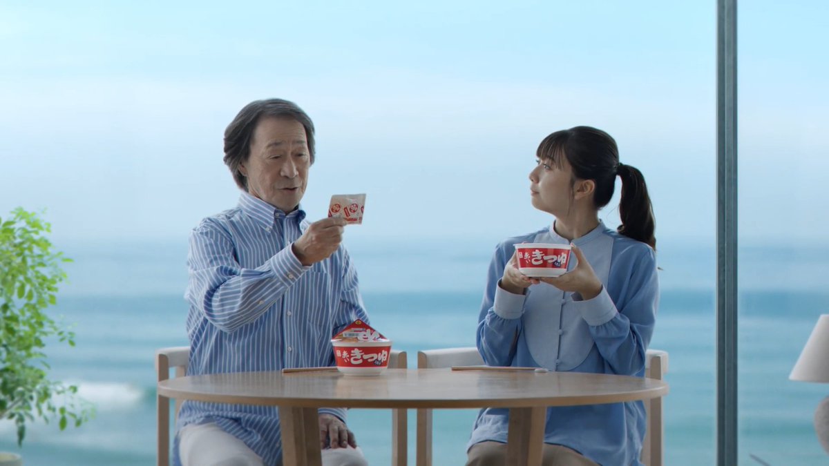 An older man and a young woman sit at a seaside table holding and examining packets of Nissin "Cup Noodles" (カップヌードル), including a red cup labeled "今 新 きつね" (Kitsune Udon). The scene depicts a new commercial featuring Tetsuya Takeda and Moe Kamishiraishi enjoying the iconic Japanese instant noodles together against an ocean backdrop.