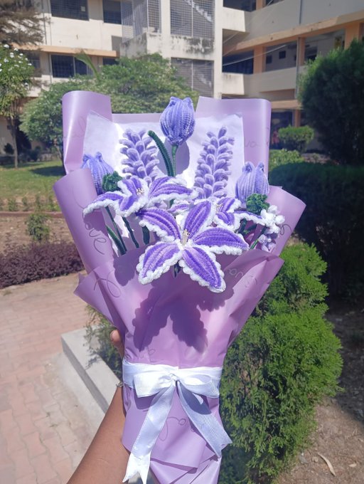 A hand holds a bouquet of crocheted purple lily flowers wrapped in light purple paper and tied with a white ribbon, set against a backdrop of a building and greenery.