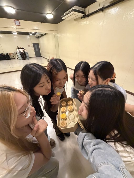 A group of six young women gathered closely together in a dance studio, looking down at a box of small pastries or cupcakes.