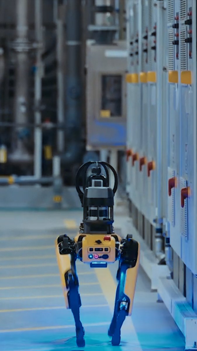 A yellow and black quadruped robot walks down an industrial corridor lined with electrical equipment panels.