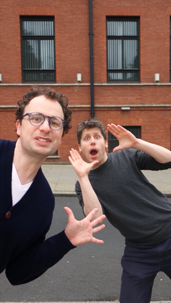 Two men making exaggerated, shocked expressions with their hands raised, posing for a selfie in front of a brick building with windows.